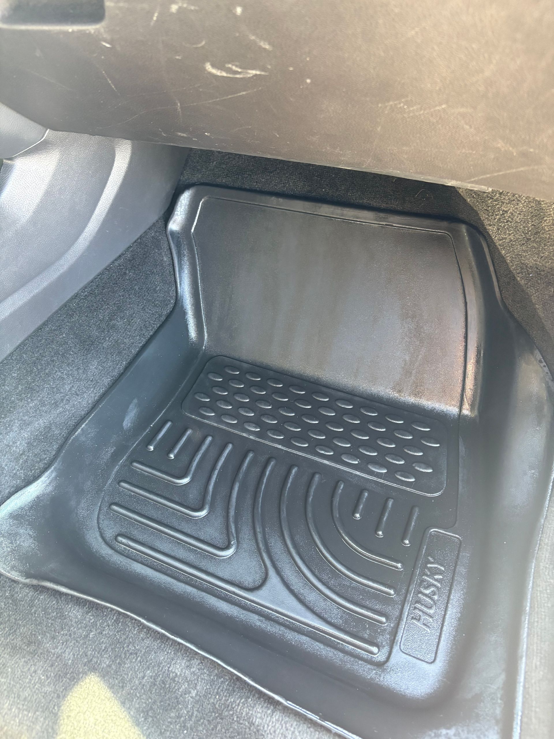 Black rubber car floor mat in a vehicle footwell, with textured grooves and raised edges.
