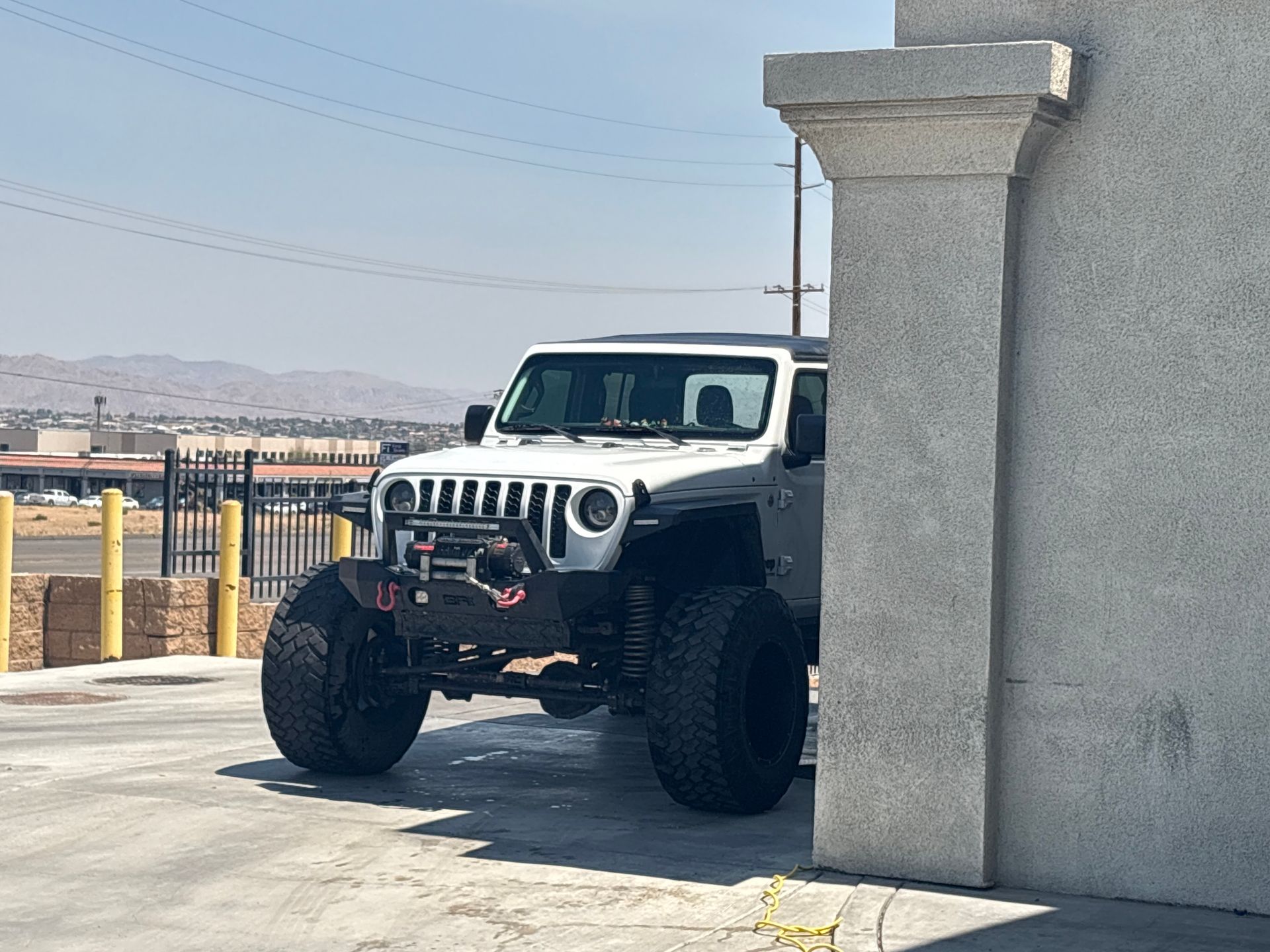 White Jeep Wrangler parked partly behind a concrete pillar in a sunny lot with mountains in the background