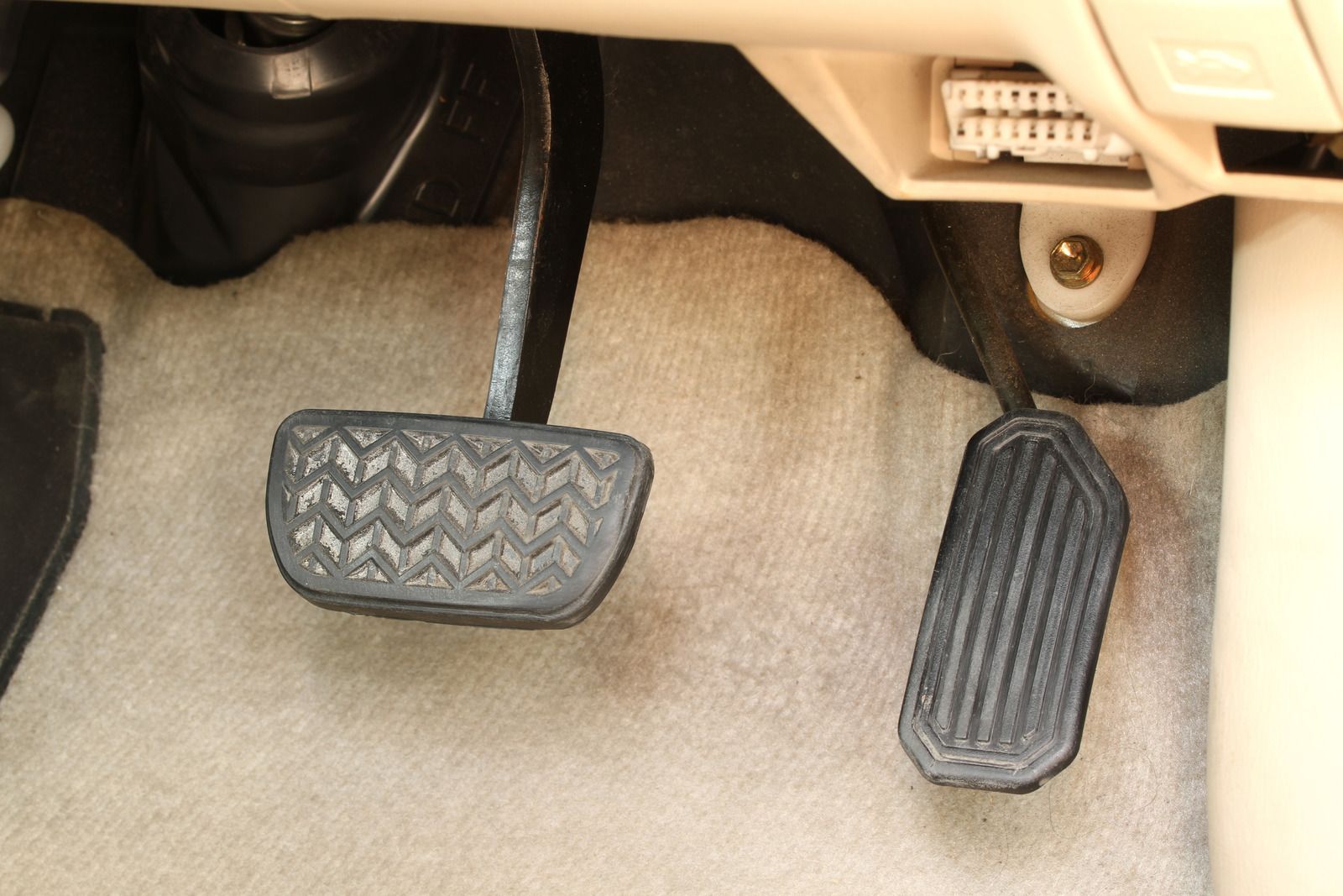 Car footwell with brake pedal, accelerator pedal, and light-colored floor mat