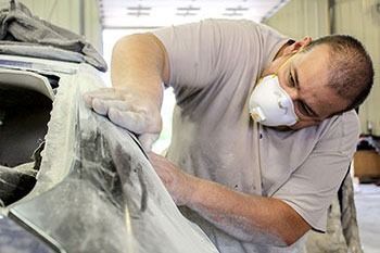 Man wearing a dust mask sands a car body in a repair shop, with white primer dust visible.