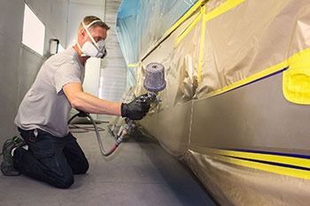 Person spray-painting a car body in a garage, wearing a mask and kneeling beside the vehicle.
