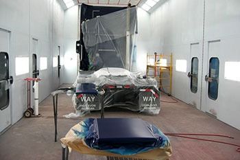 Car wrapped in plastic inside a spray booth, with a blue part on a stand in the foreground