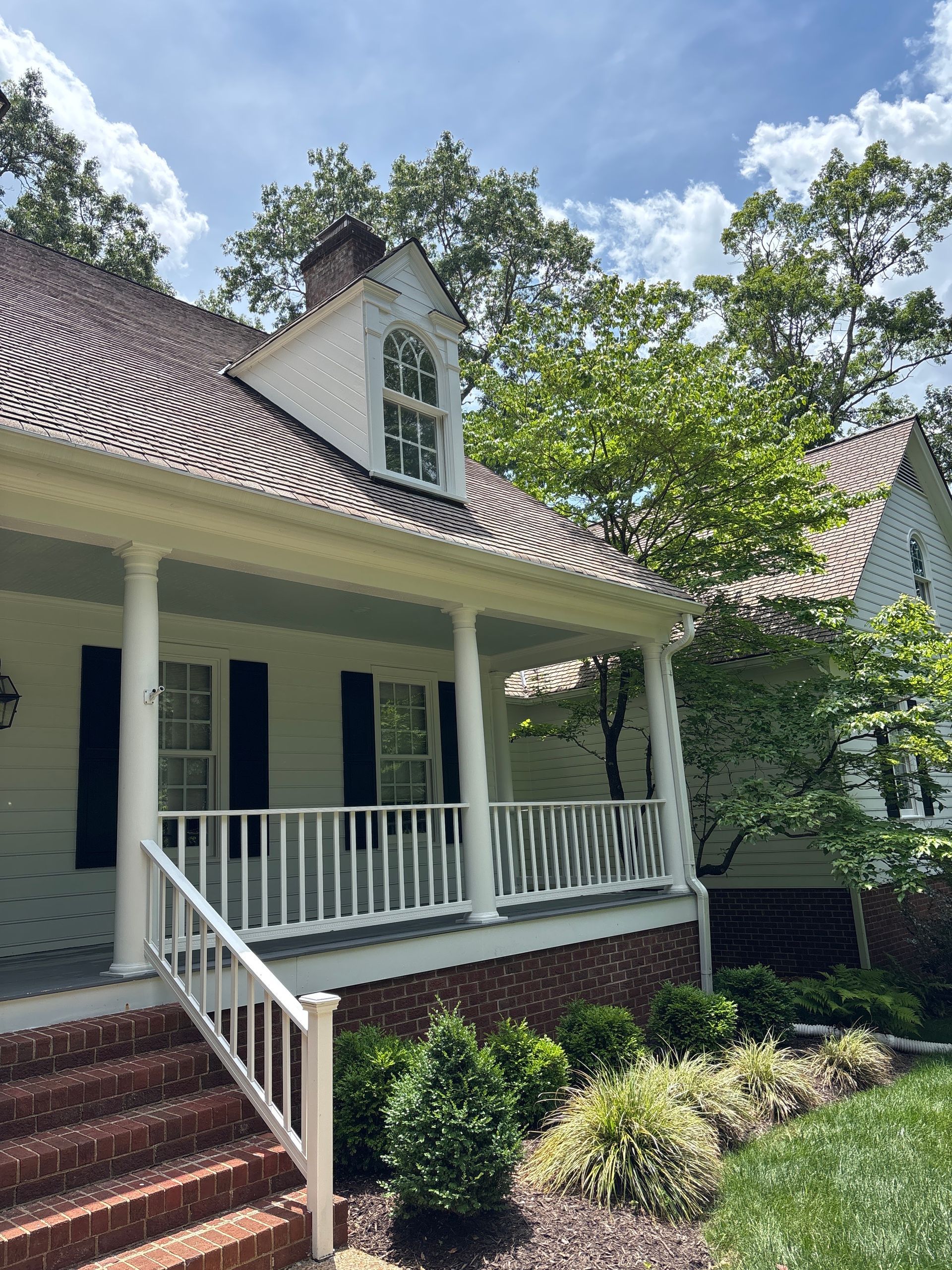 White house with steep shingle roof, front porch, brick steps, and lush green yard under a blue sky