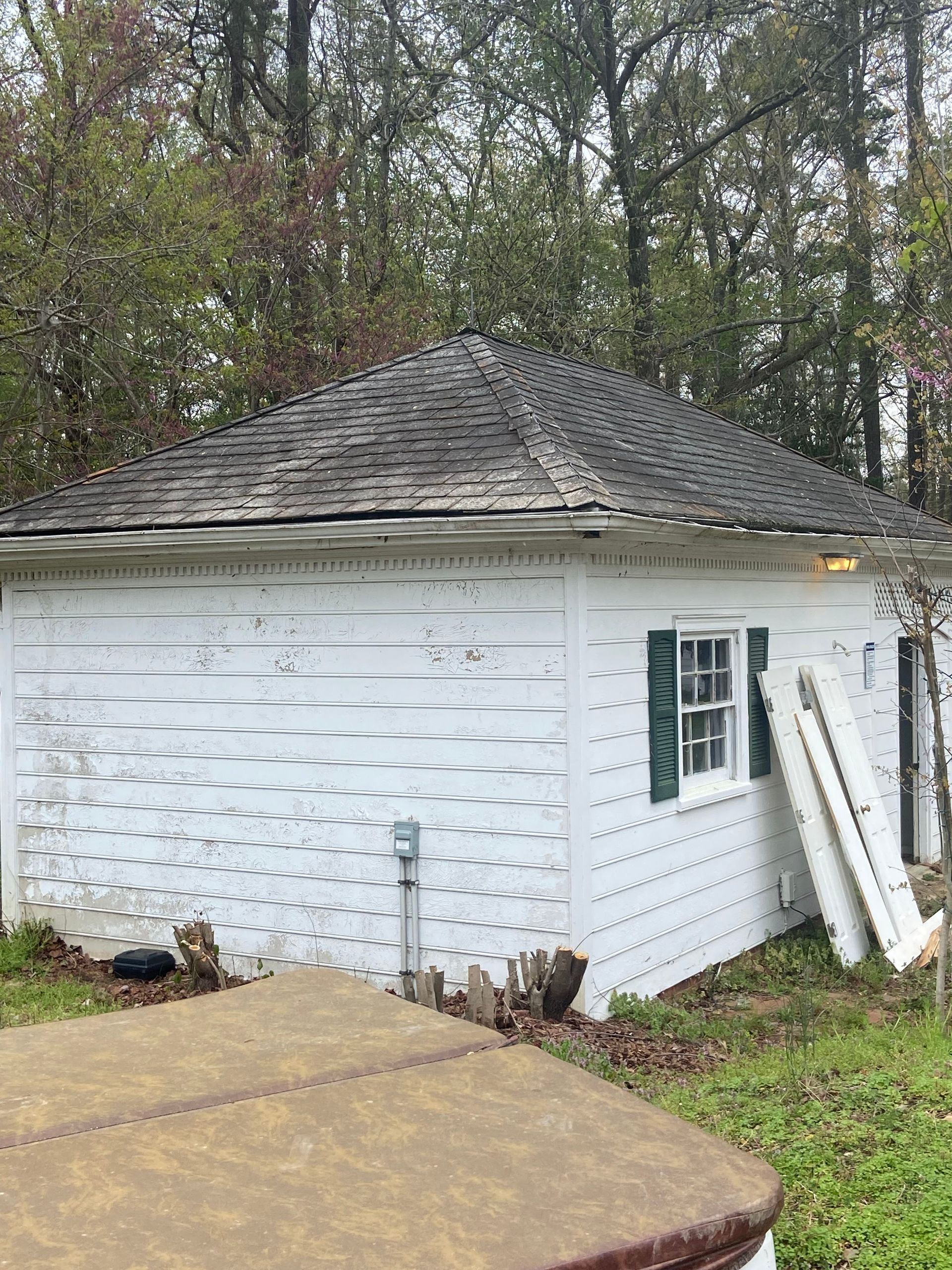 White garage with damaged siding, dark shingled roof, and leaning boards beside a concrete driveway.