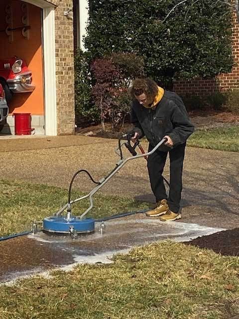 Person pressure-washes a driveway with a blue surface cleaner near a garage and lawn.