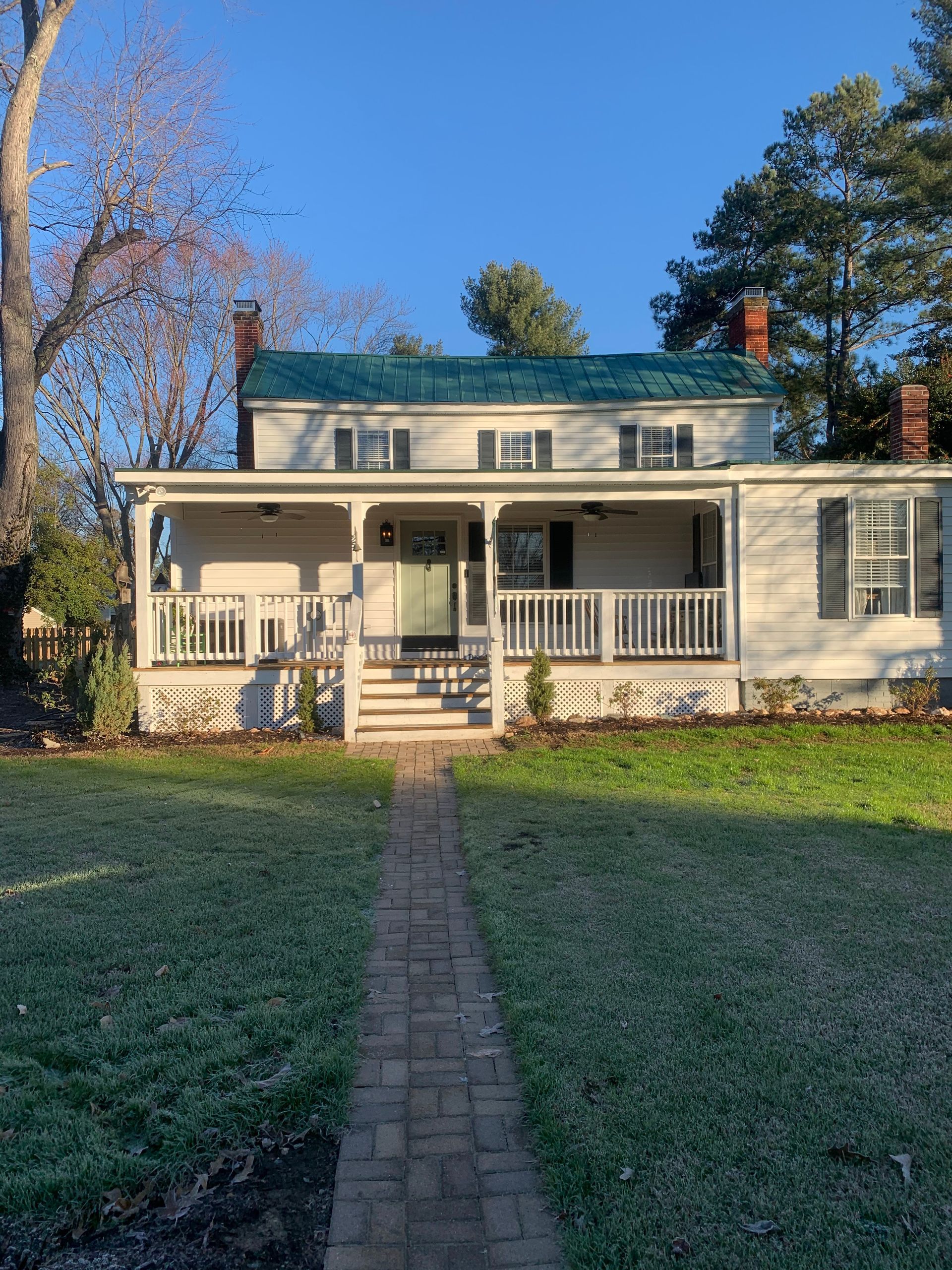 Small house with white porch and green roof, framed by a brick walkway and grassy yard.