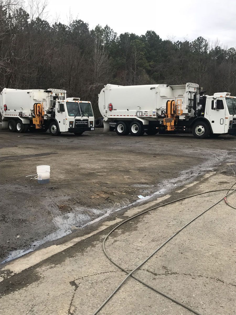 Two white garbage trucks parked on a muddy lot near a wooded tree line