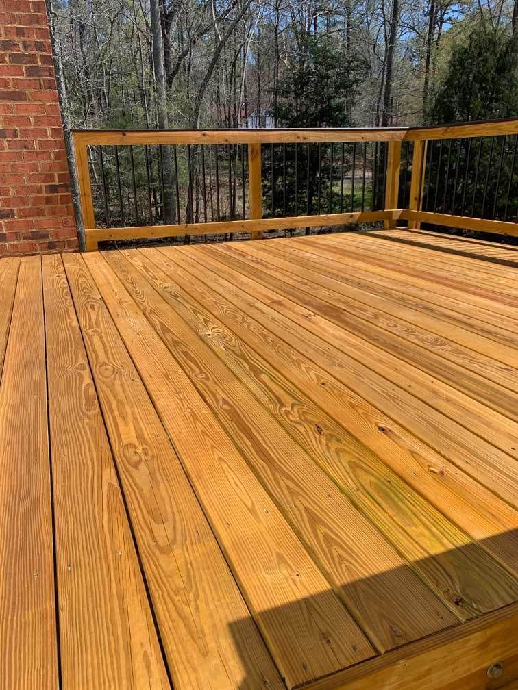 Wooden deck with railing beside a brick wall, overlooking trees in the background.