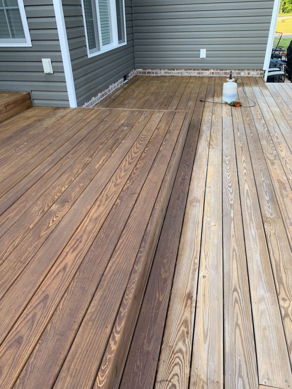 Weathered wooden deck boards beside gray siding, showing two tones of wood after partial replacement.