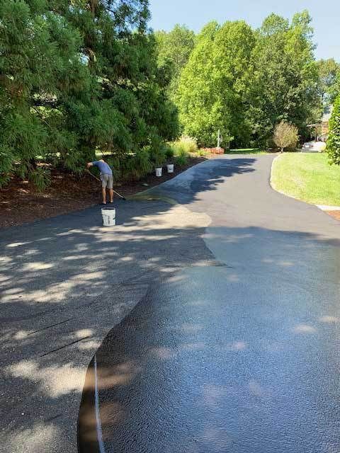 Workers painting a curved asphalt driveway beside trees and shrubs on a sunny day