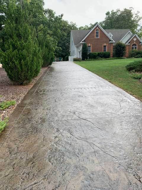 Wet concrete driveway leading to a brick house with trees and lawn on a cloudy day