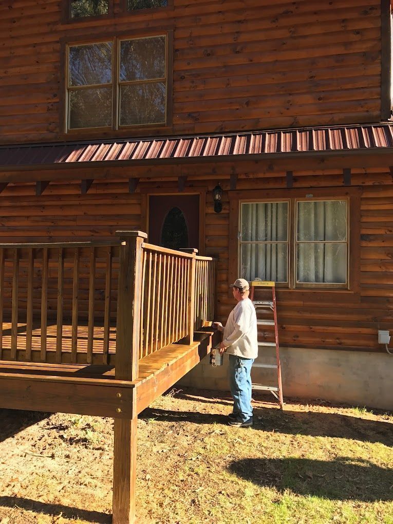 Person standing beside a wooden cabin porch in sunlight.