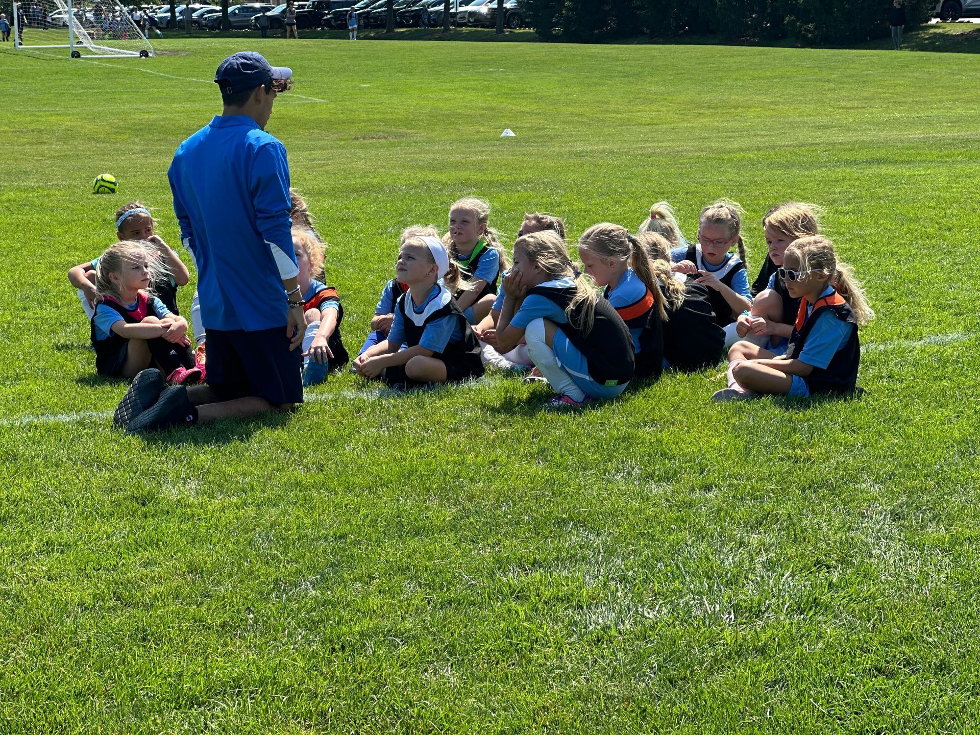 A group of young girls are sitting on the grass while a man talks to them.
