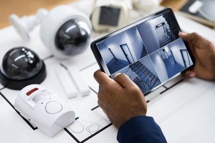 Person holding a tablet displaying security camera feeds, surrounded by security equipment in blue tones.