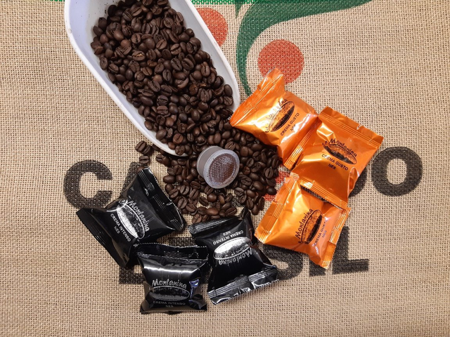 Un cucchiaio che versa chicchi di caffè su un sacco di iuta, accanto a cioccolatini incartati in carta nera e arancione e una singola capsula di caffè.