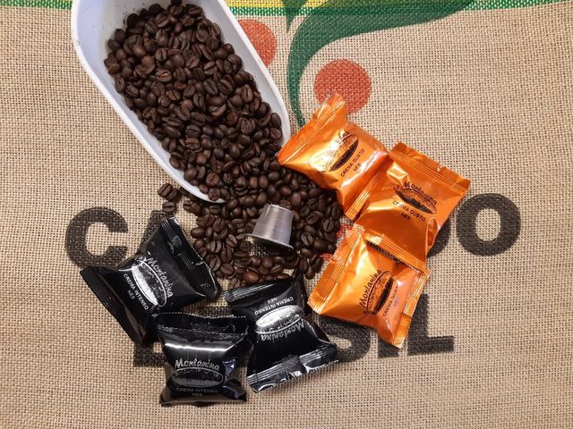 Chicchi di caffè tostati che vengono versati da un mestolo su un sacco di iuta, con capsule di caffè nere e arancioni sparse qua e là.