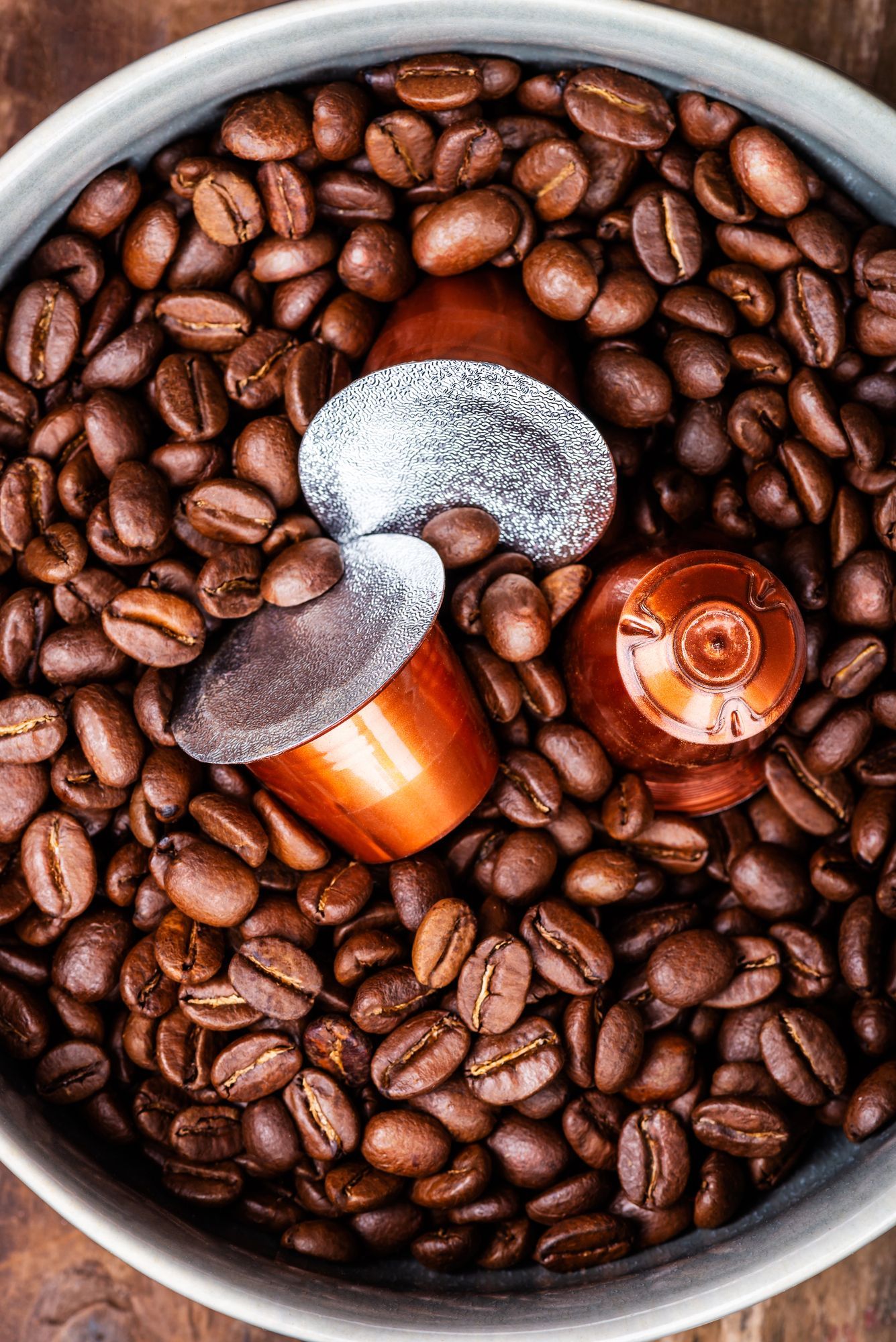 Una ciotola piena di chicchi di caffè tostati e tre capsule di caffè metalliche.