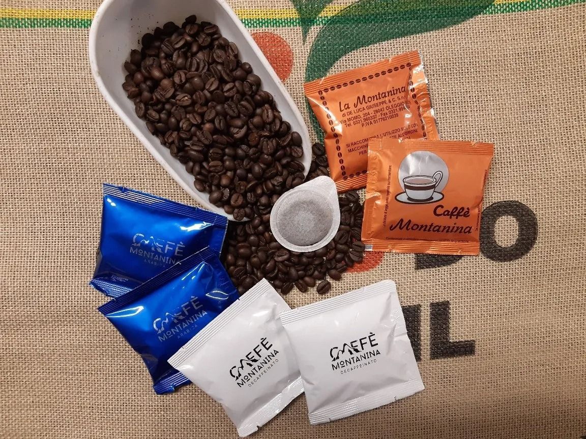 Chicchi di caffè in un misurino con bustine individuali di caffè blu, arancioni e bianche su uno sfondo di tela di iuta.