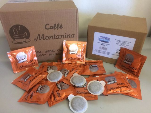 Due scatole di cartone con la scritta "Caffè Montanina" circondate da numerose capsule di caffè arancioni su una superficie bianca.