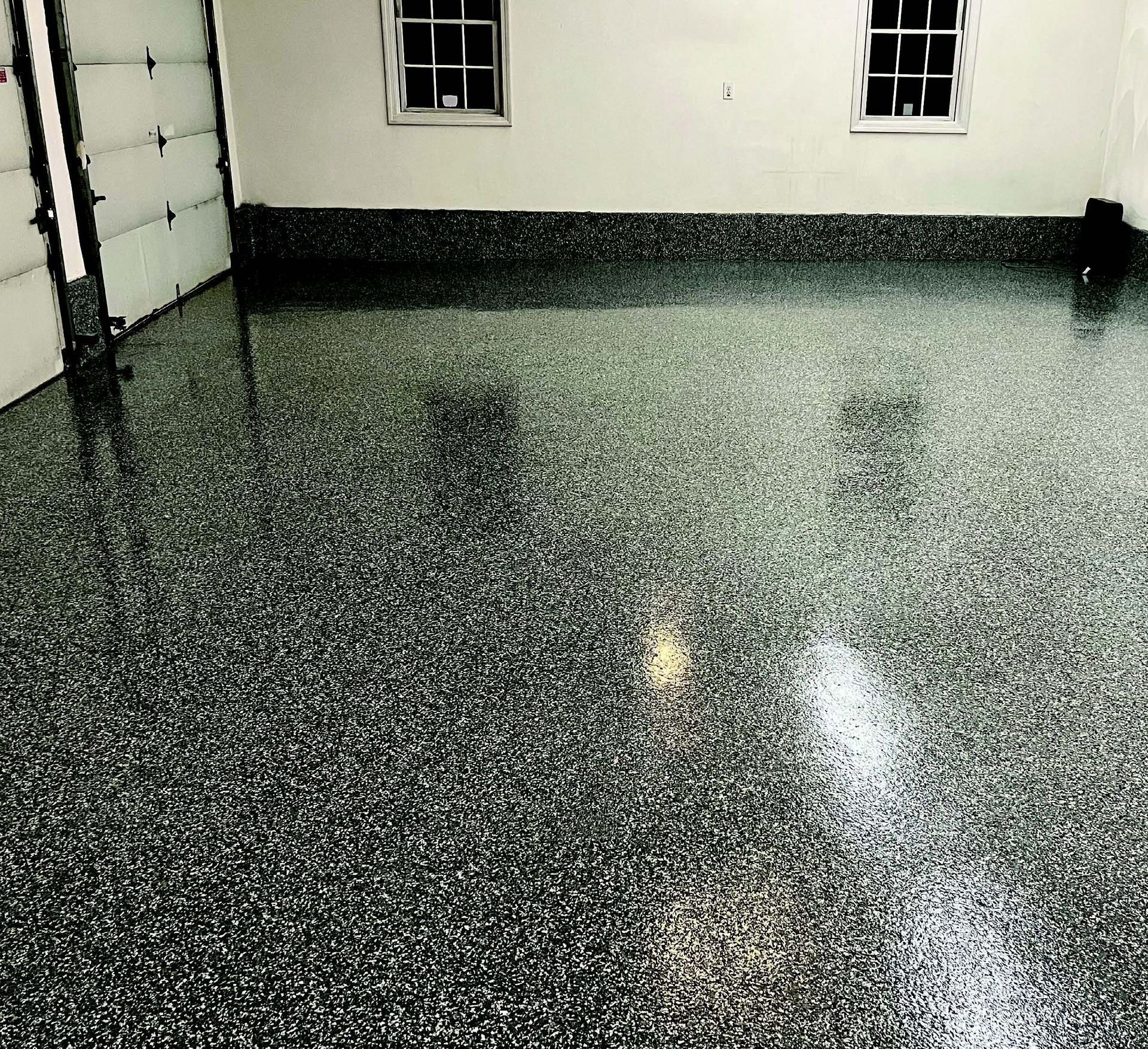Empty garage with a glossy black-speckled floor and white walls, reflecting light from windows.