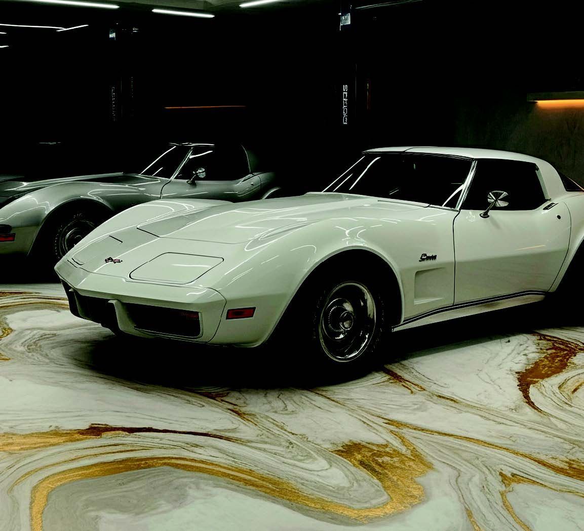 White classic sports car in a showroom, with a dark convertible behind it on a patterned floor.