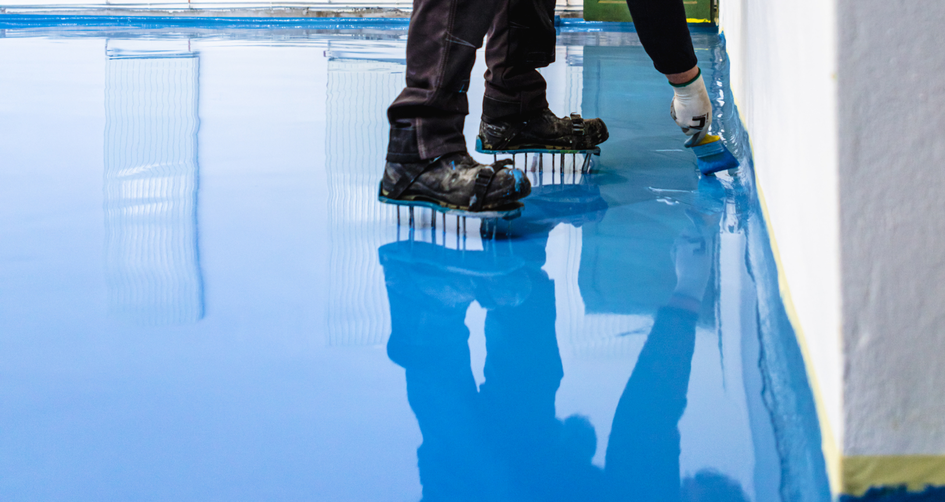 Workers applying a glossy blue floor coating with rollers in a room.