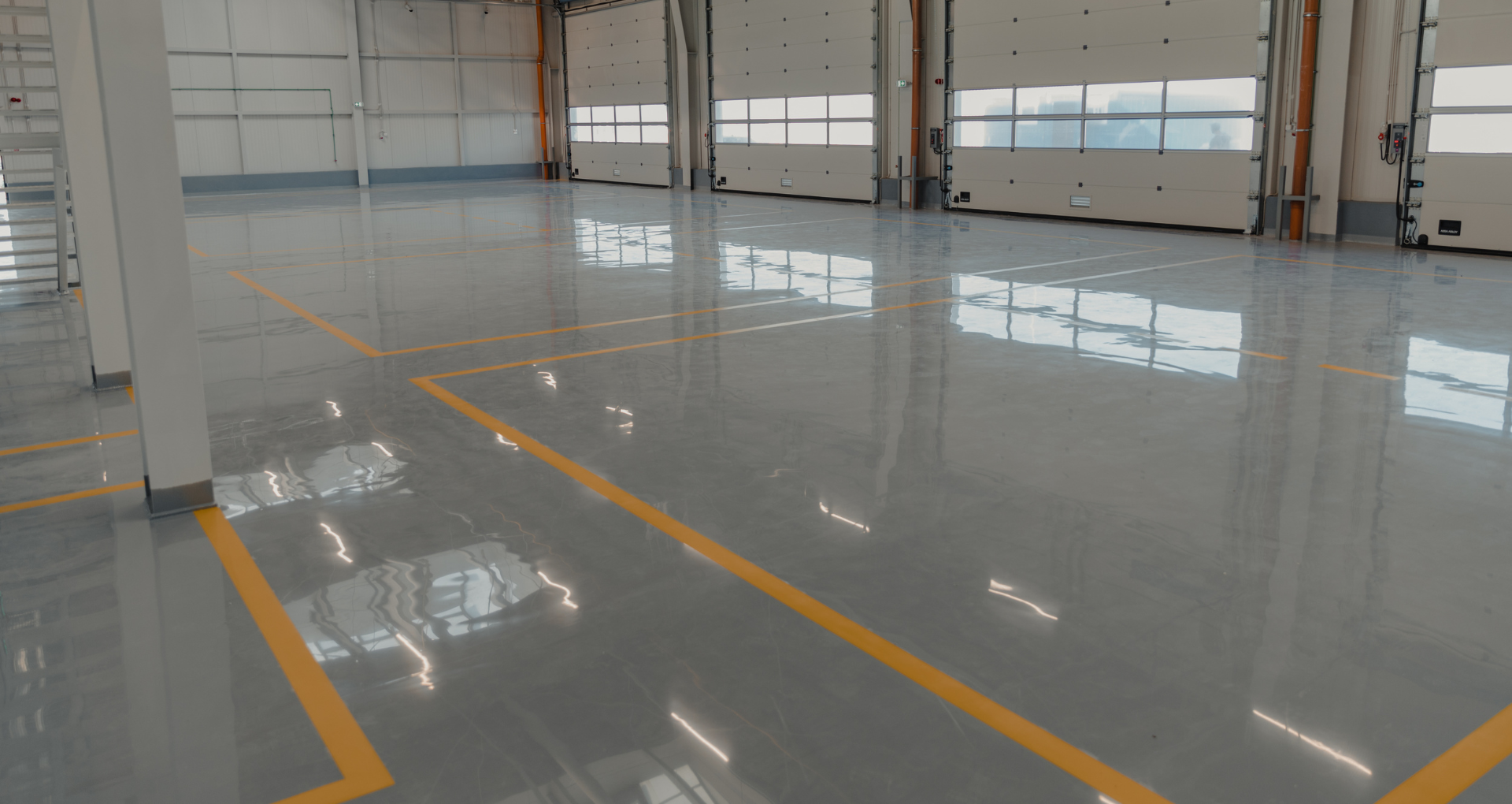 Empty industrial warehouse with glossy gray floor and yellow lane markings, large open bay doors in back.