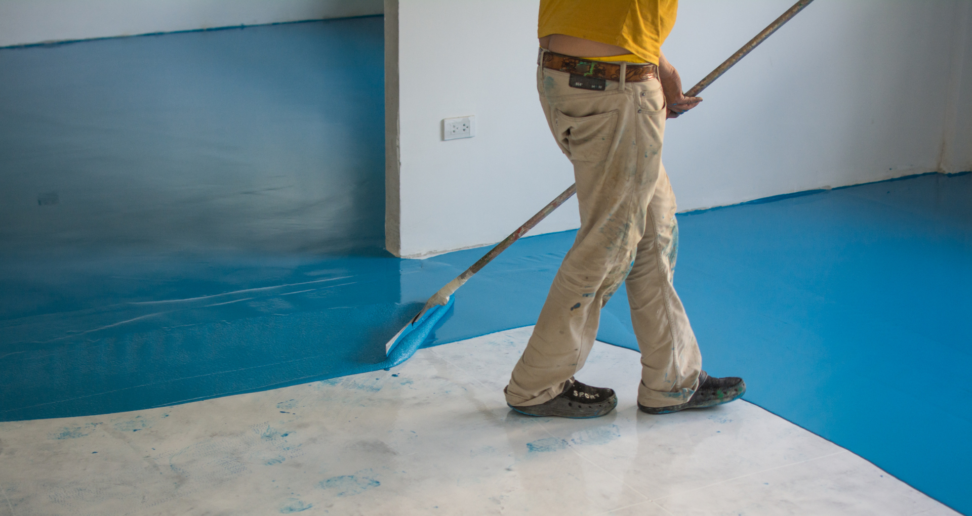 Person spreading blue floor coating with a long-handled tool in a room under renovation