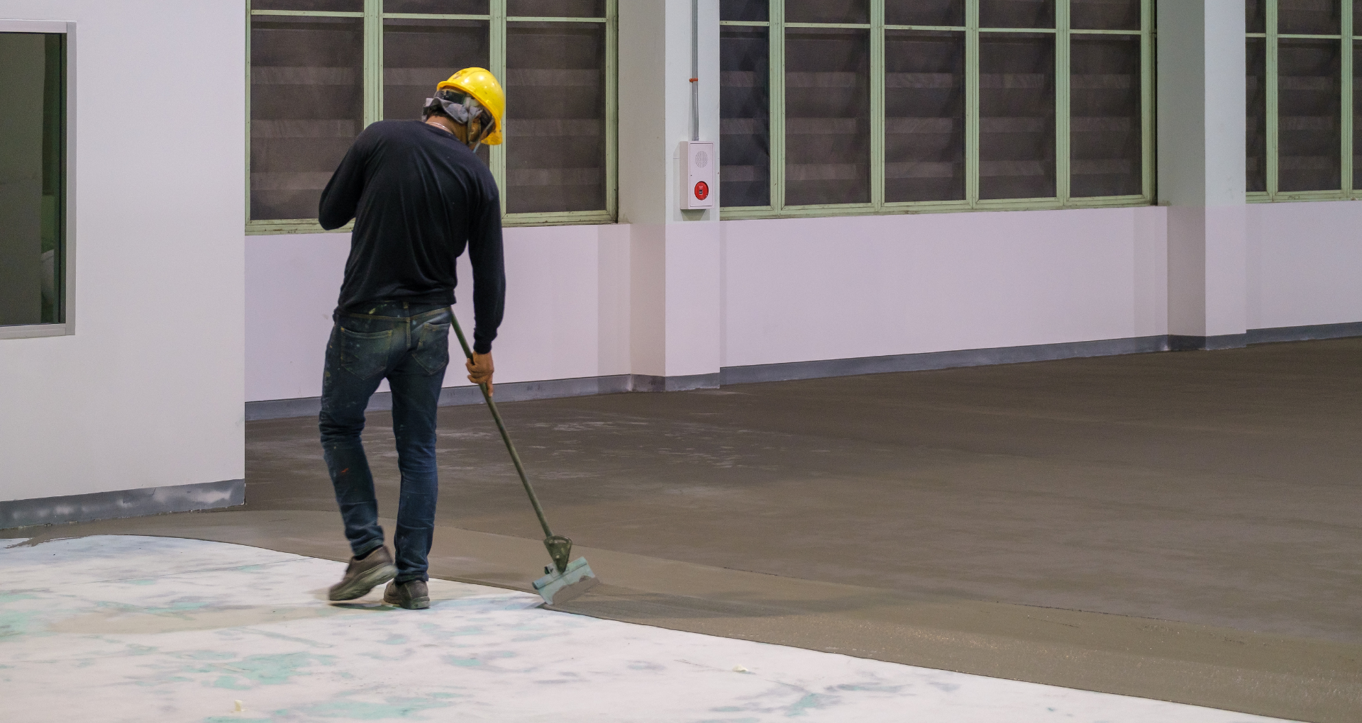 Worker in a yellow hard hat smoothing a wet concrete floor outside a building.