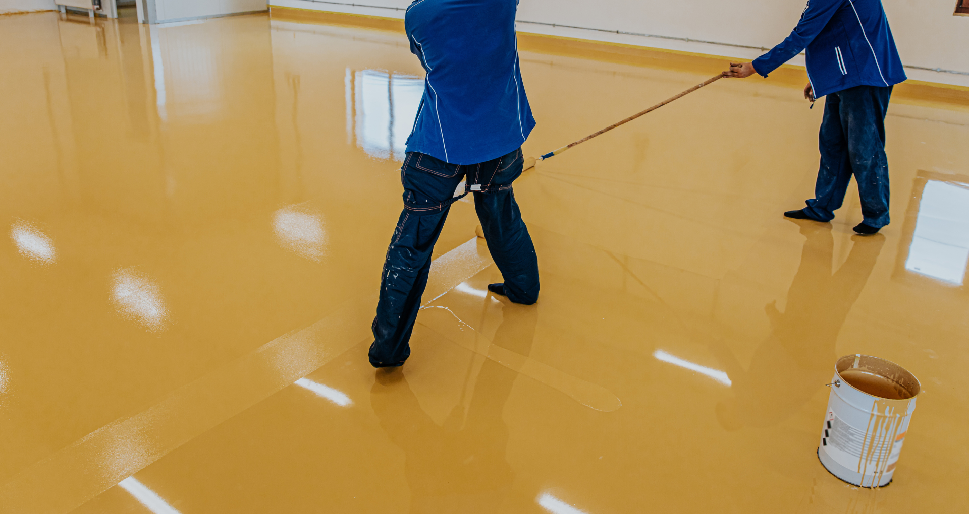 Two people spreading glossy yellow floor coating in a room, with a paint bucket on the floor.