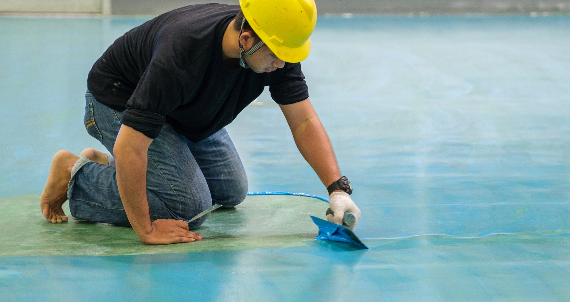 Worker in a yellow hard hat kneels on a blue floor, applying coating with a trowel.