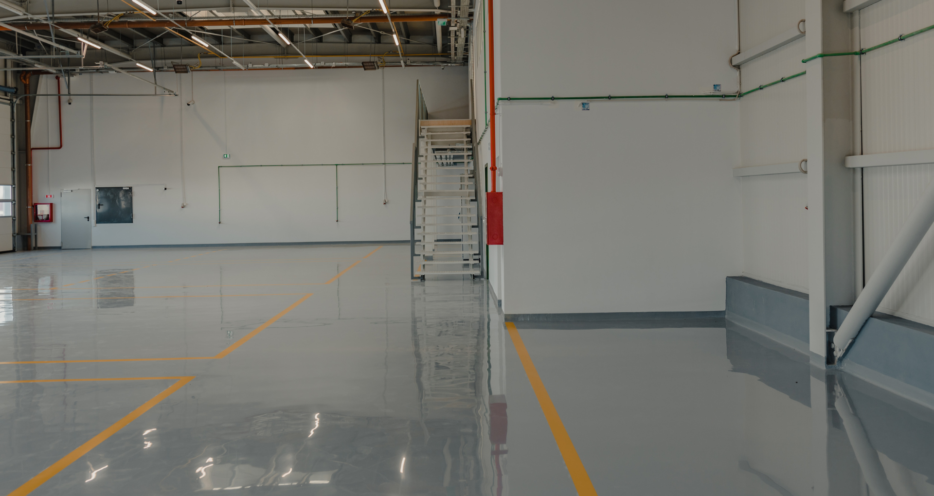 Empty industrial warehouse interior with polished gray floor, white walls, exposed ceiling beams, and yellow floor markings