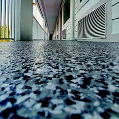 Low-angle view down a building corridor with speckled floor, columns, and louvered vents on the right.