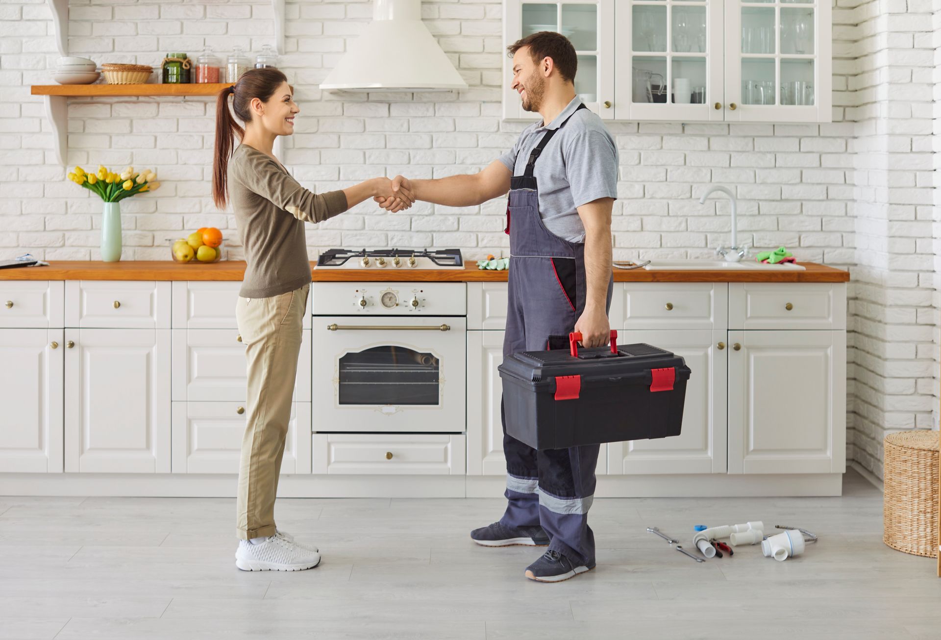 A woman knells beside a drain leaking water into a bucket, holding her cell phone. Plumber fixing a kitchen sink under a white cabinet, with tools on the counter.
