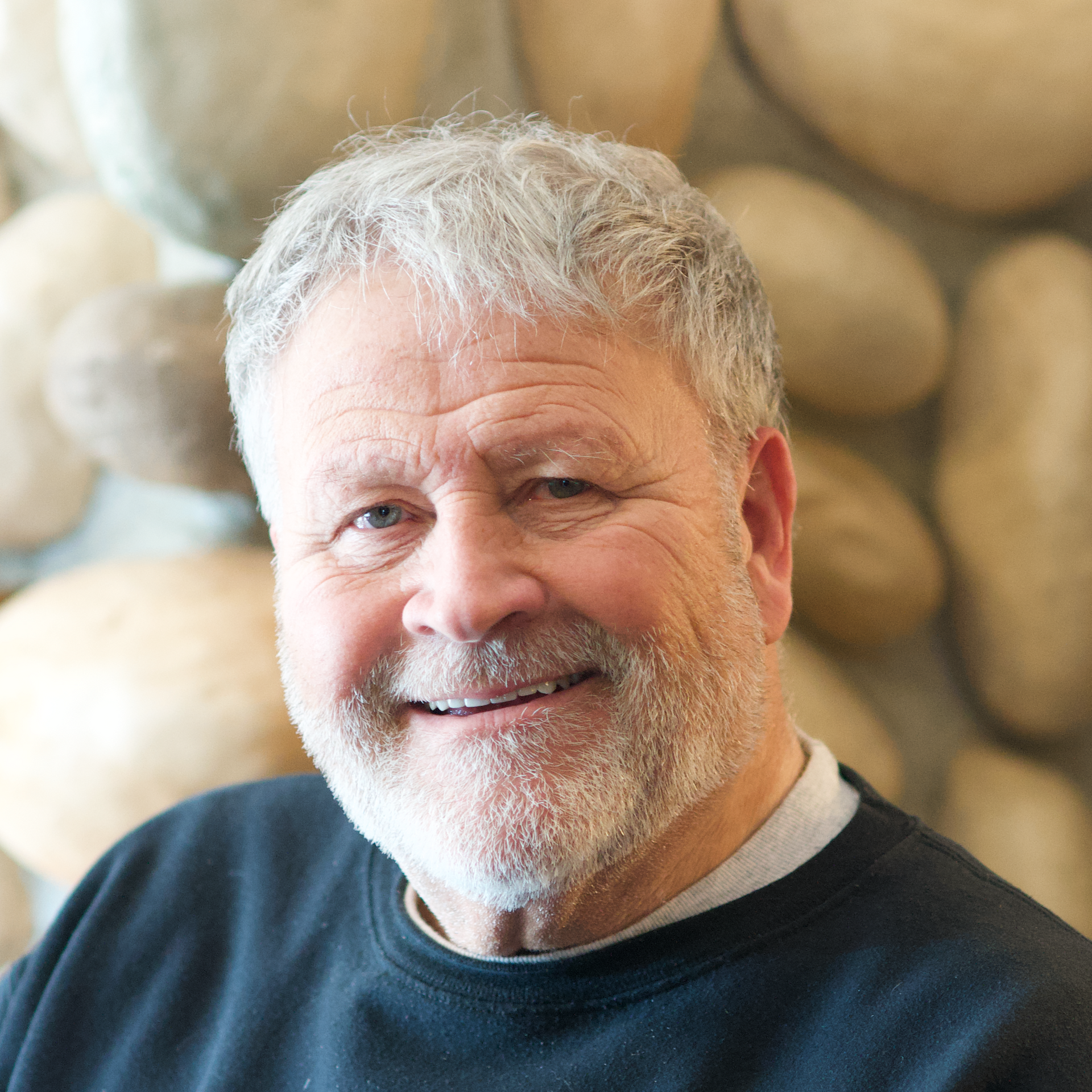 Smiling man with gray hair and beard in a dark sweater against a stone wall background