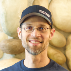 Smiling person wearing glasses and a dark cap, standing in front of a stone wall.