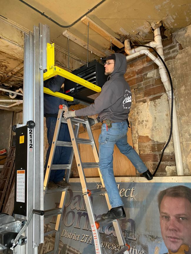 Worker in a basement installing a yellow lift, with a ladder and exposed pipes nearby.