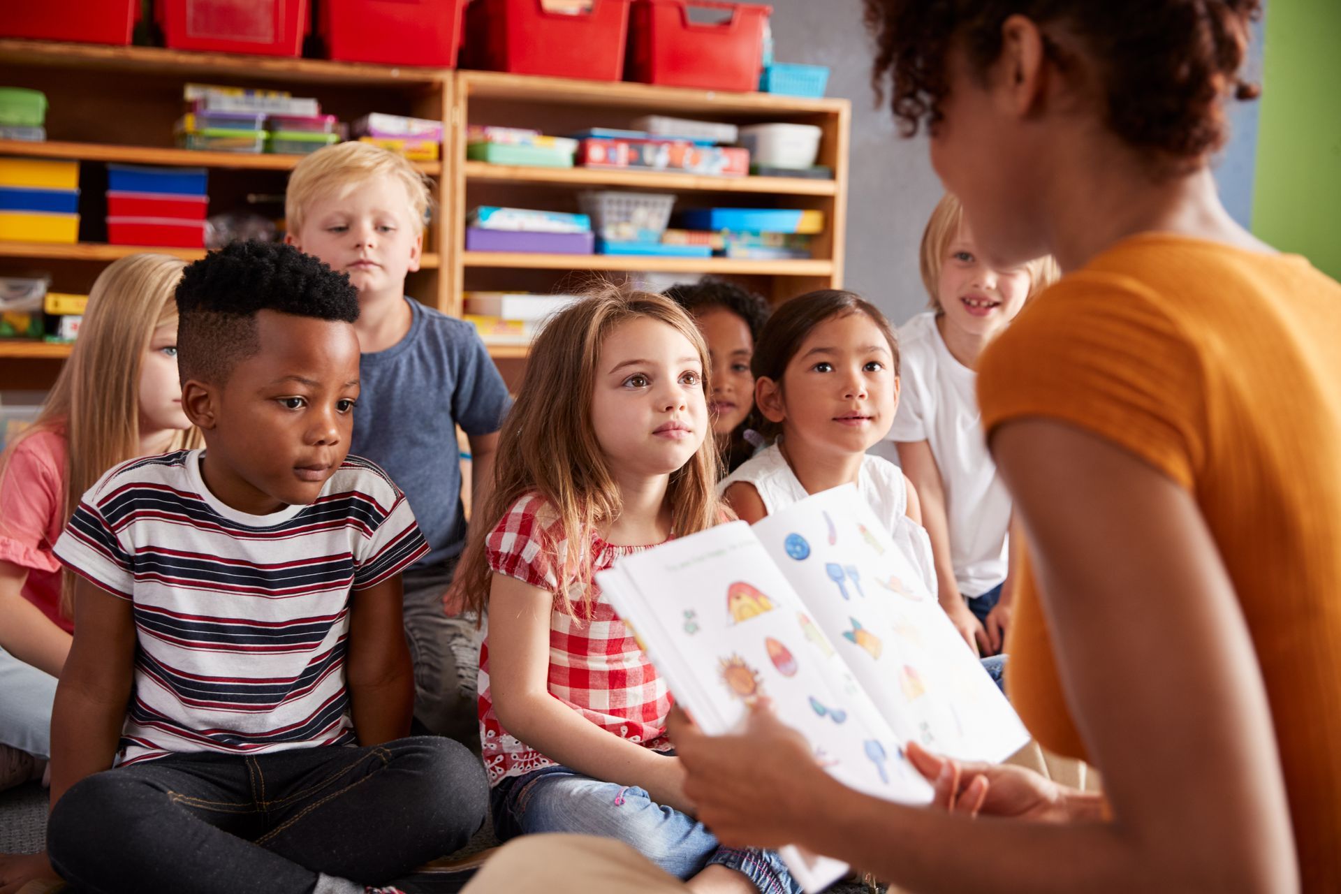 A woman reading a storybook to a group of young children sitting on the floor in a colorful room. A woman reading a storybook to a group of young children sitting on the floor in a colorful room.
