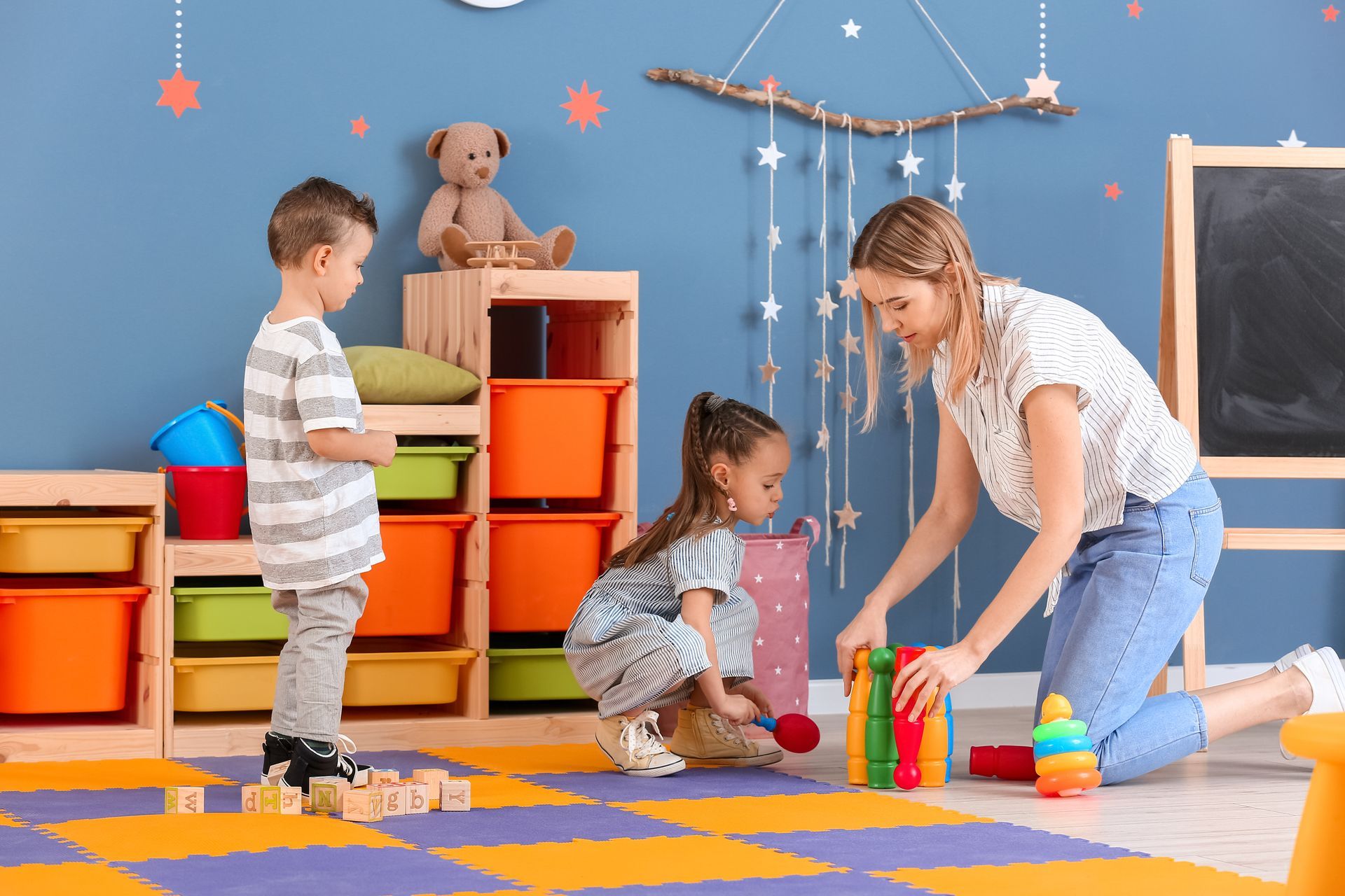 An adult smiles as two children draw and play at a small round table. A teacher and two children playing with colorful toys in a daycare classroom.