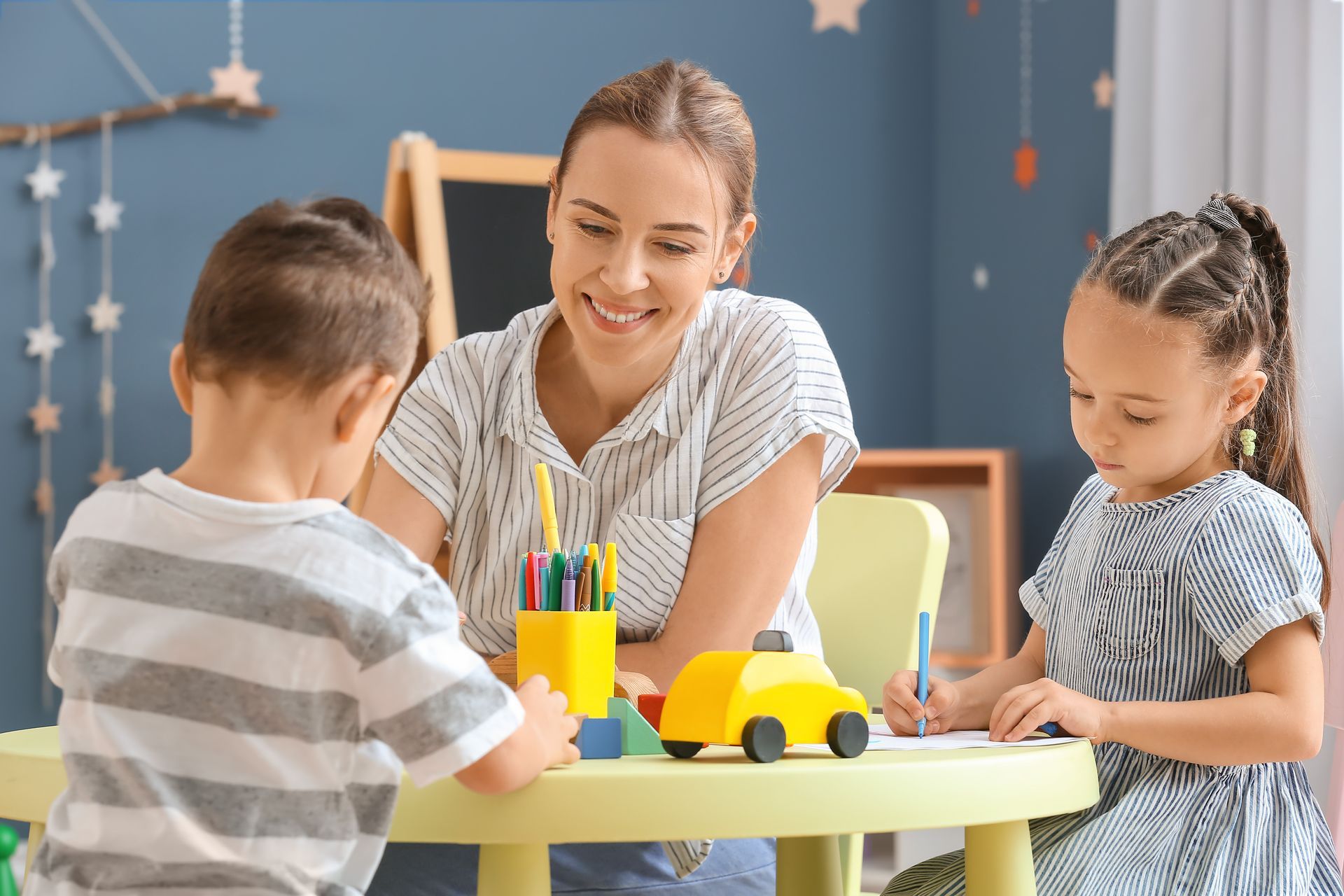 An adult smiles as two children draw and play at a small round table.