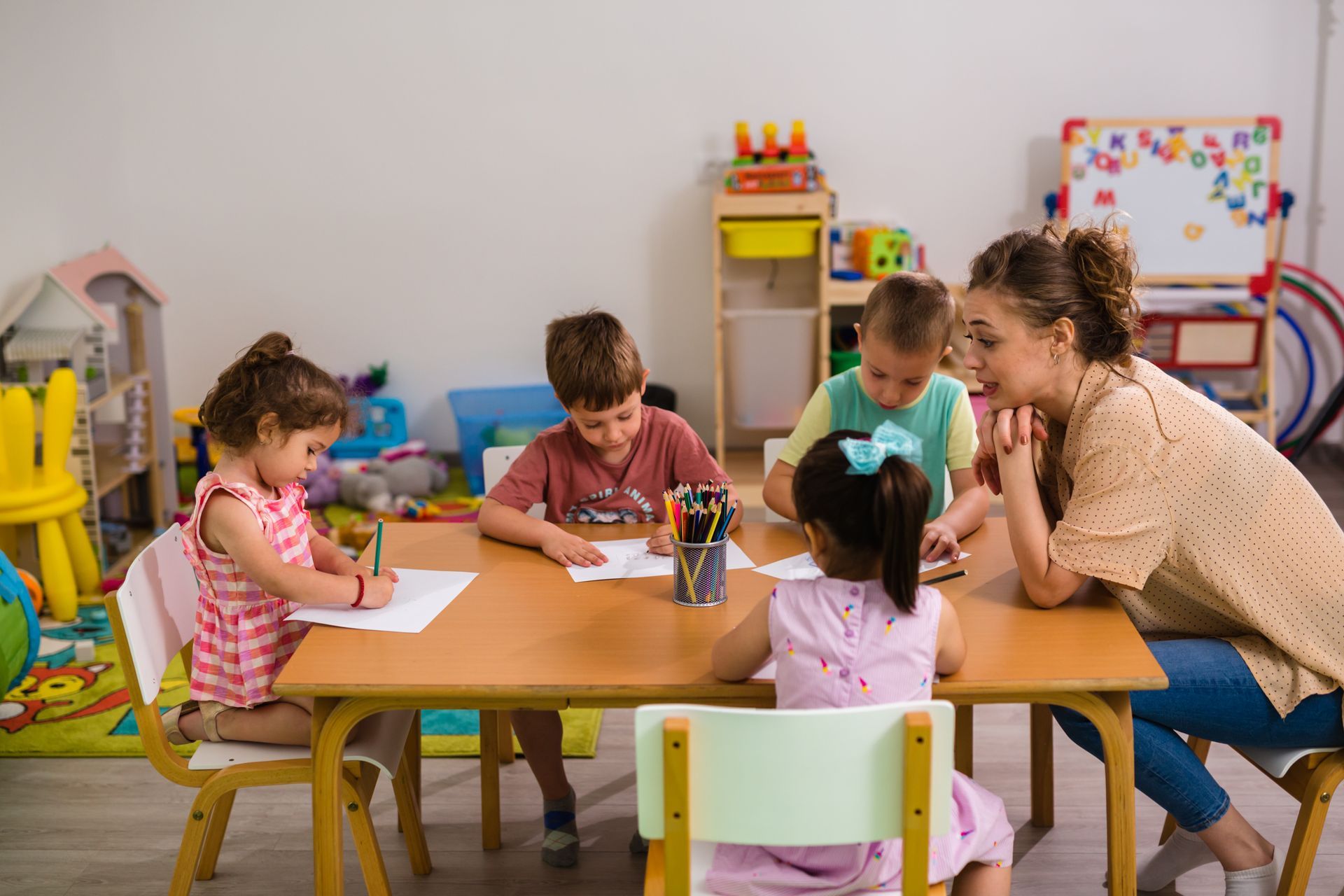 Teacher helps four children draw at a table in a child daycare agency classroom. Teacher helps four children draw at a table in a child daycare agency classroom.