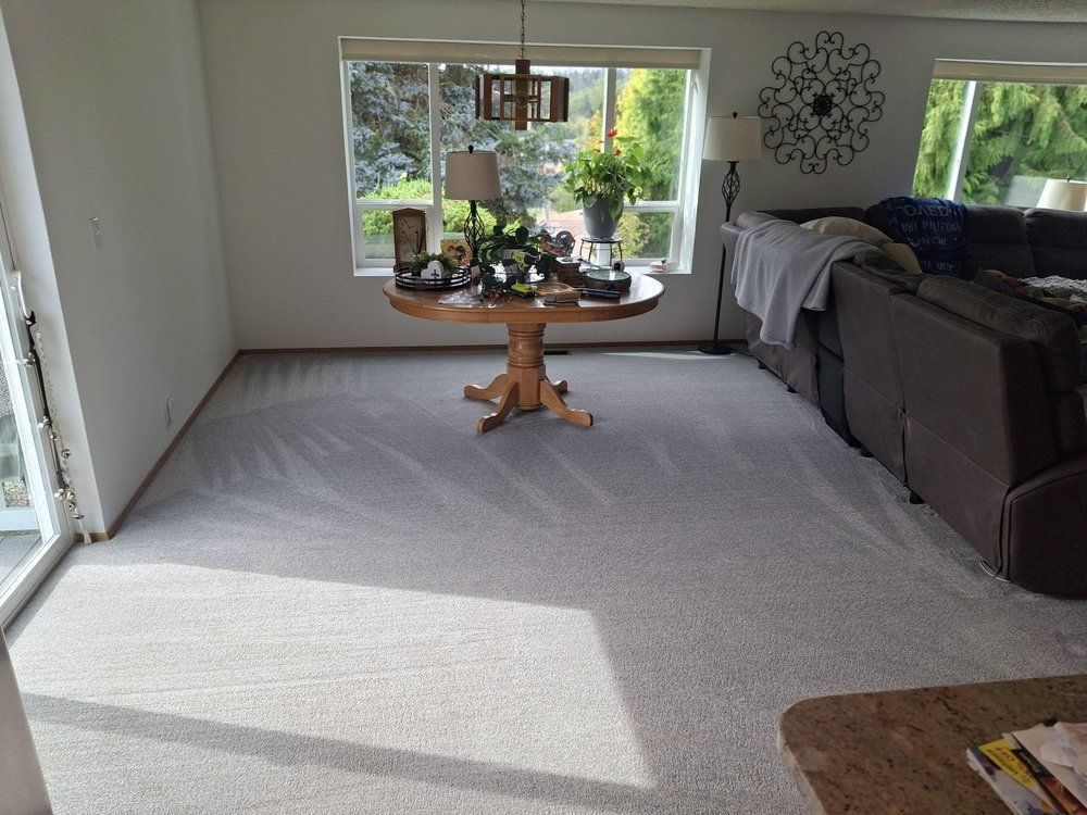 Bright living room with gray carpet, dark sofa, and a round wooden table by a window.
