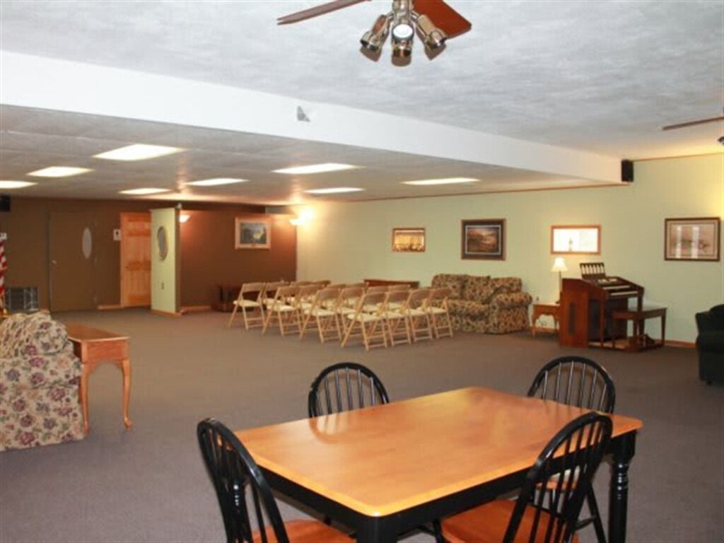 Large room with chairs, tables, and couches arranged for a gathering; beige walls, brown accents.