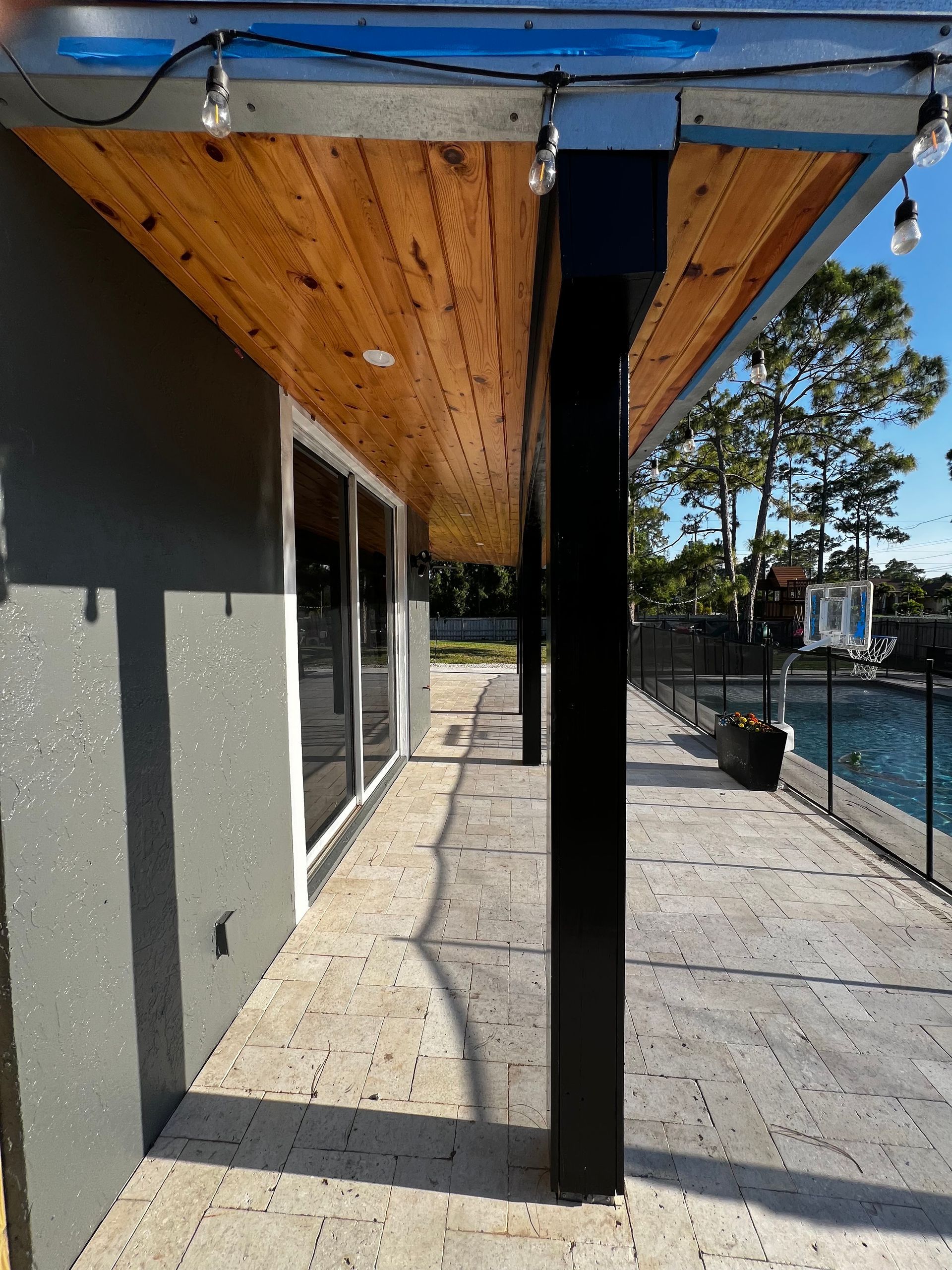 Covered outdoor walkway beside a gray building, with wood soffit, black post, and fence by water.