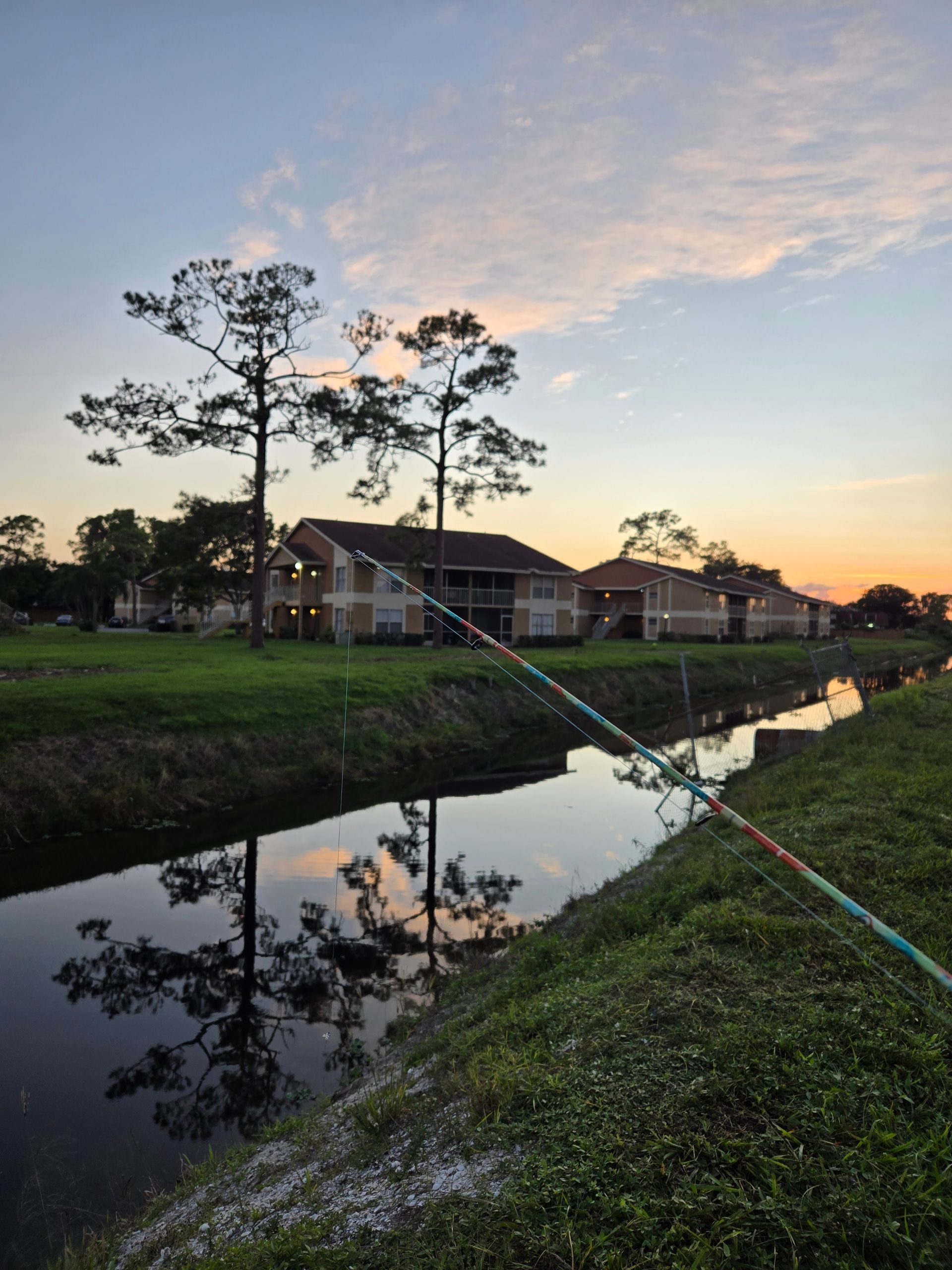 Sunset over a canal reflecting houses, trees, and a colorful sky along a grassy bank.