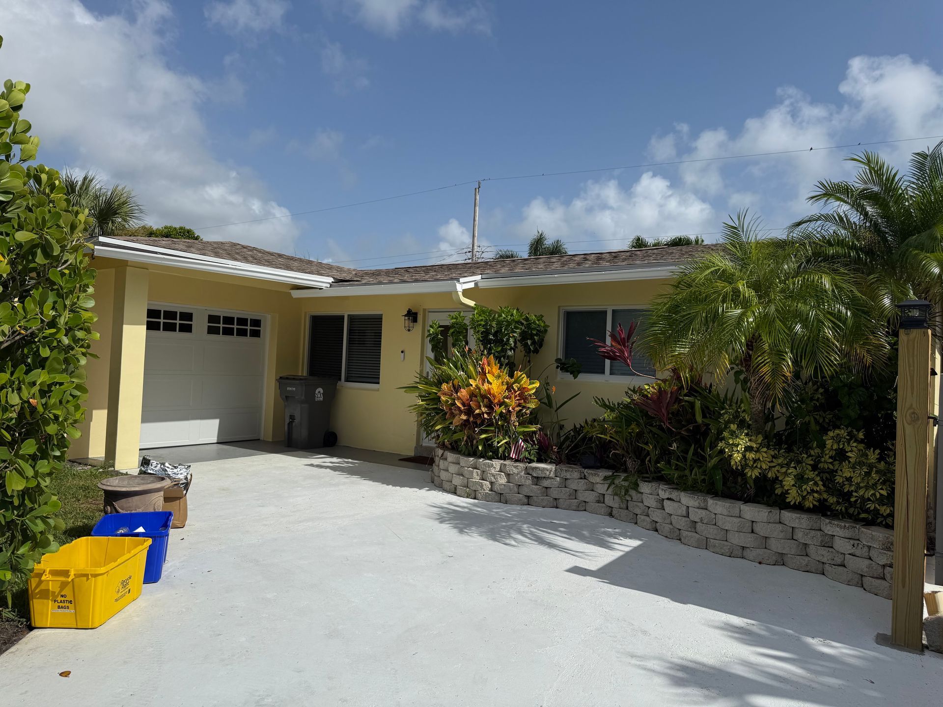 Single-story house with a driveway, colorful bins, and tropical plants under a sunny blue sky