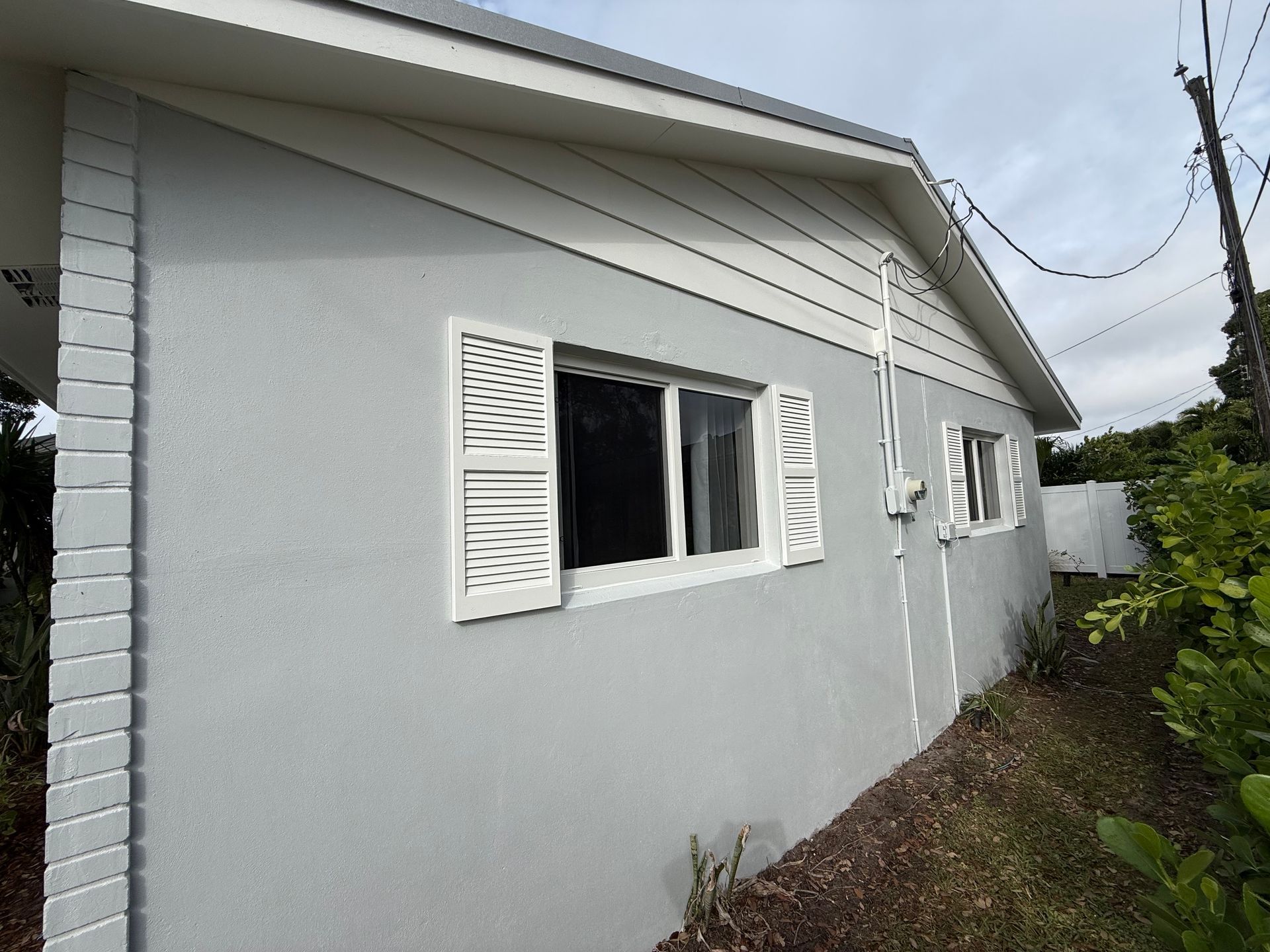 Side view of a light gray house with white shutters and windows along a narrow yard
