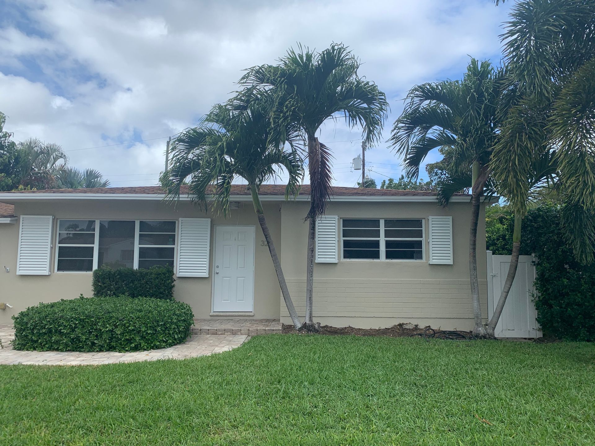 Single-story beige house with white shutters, palm trees, and a green lawn under a cloudy sky