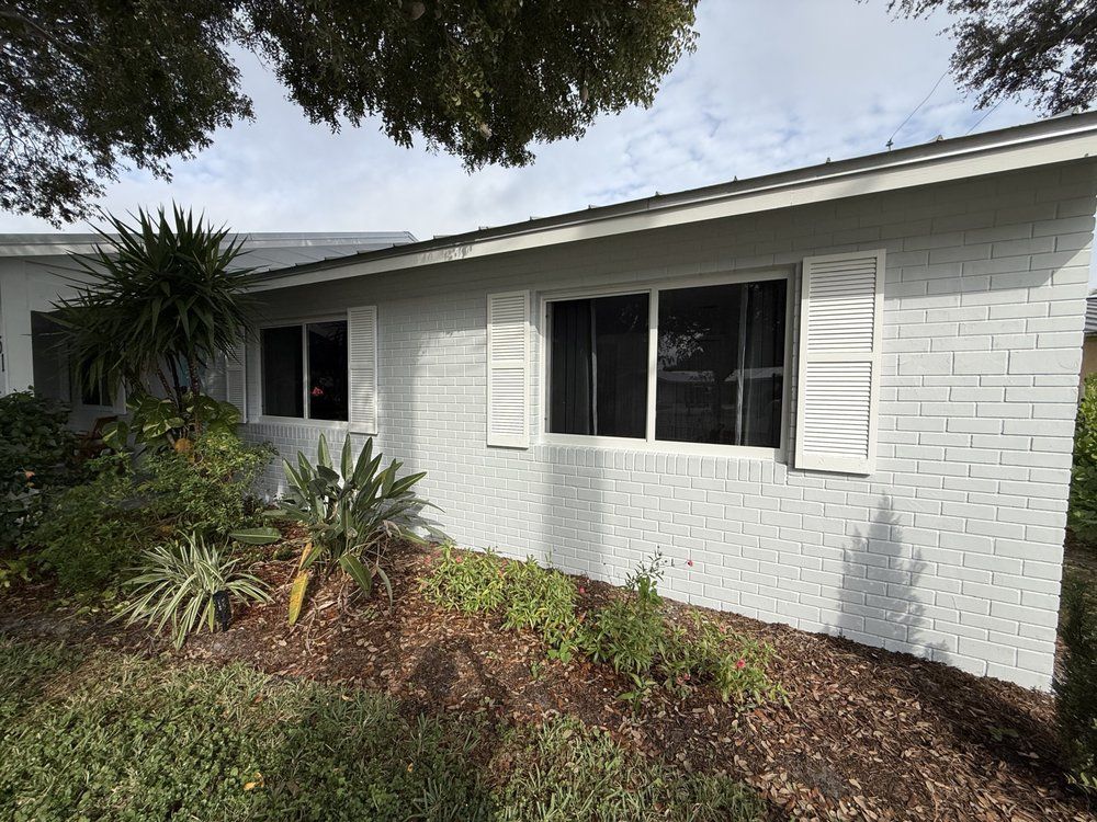Single-story white brick house exterior with dark windows, shutters, and landscaped front garden