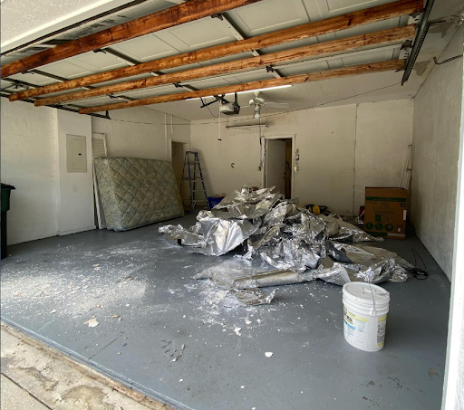 Garage interior with unfinished drywall debris, exposed ceiling beams, and a white bucket on the floor